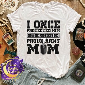 Puede incluir: Camiseta blanca con texto negro que dice "I once protected him now he protects me proud army mom". El texto est&aacute; rodeado de un contorno negro. Una placa de identificaci&oacute;n plateada con una peque&ntilde;a bandera estadounidense cuelga del texto.