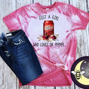 May include: Pink bleached t-shirt with a white graphic of a Dr Pepper can and flowers. The text "Just a girl who loves Dr Pepper" is printed below the can.