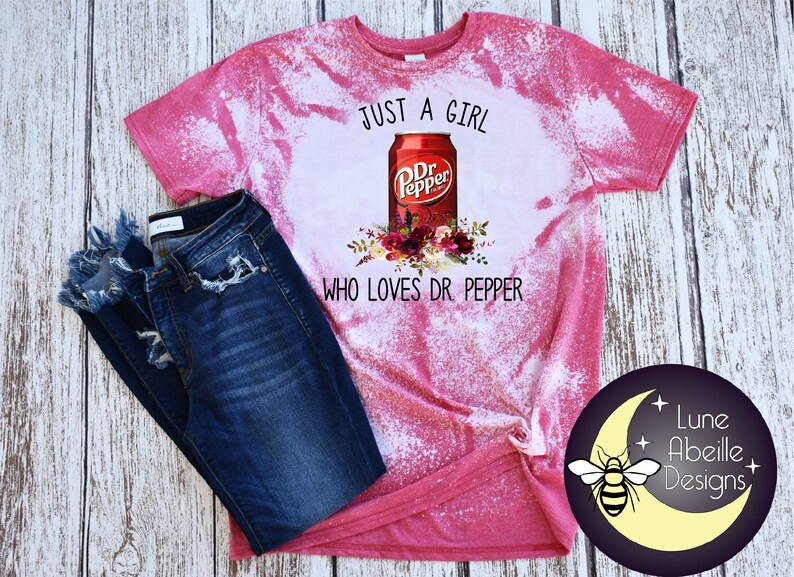 May include: Pink bleached t-shirt with a white graphic of a Dr Pepper can and flowers. The text "Just a girl who loves Dr Pepper" is printed below the can.