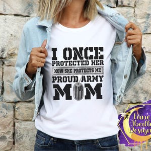 Puede incluir: Camiseta blanca con texto negro que dice "I once protected her now she protects me proud army mom" con un peque&ntilde;o gr&aacute;fico de la bandera estadounidense.