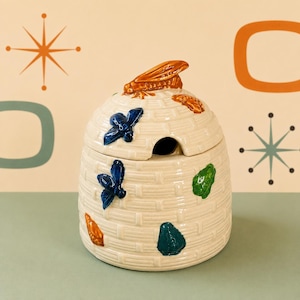 May include: A cream-colored ceramic honey pot shaped like a beehive with a woven texture. The lid features a sculpted bee handle, and the sides are decorated with small, raised, glossy bees in blue, green, and orange colors throughout the exterior.
