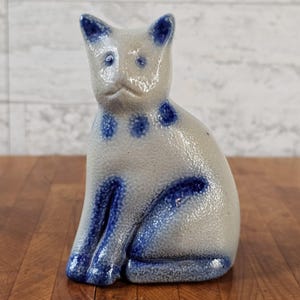 May include: A ceramic cat figurine with a speckled light gray finish and vibrant blue painted accents on the ears, eyes, neck, and legs. The cat is in a seated position with a smooth, glossy glaze covering the entire surface of the piece.