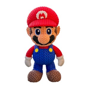 May include: A handmade, knitted Mario figure. The figure features a red hat with a white "M", a red shirt, blue overalls with yellow buttons, and brown shoes. The figure has a black mustache and blue eyes.