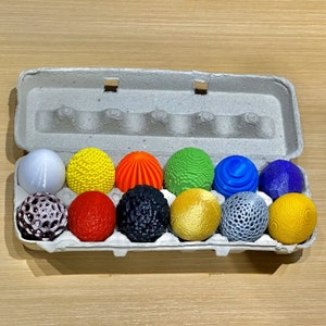 May include: A set of 12 colourful, patterned Easter eggs in a cardboard egg carton. The eggs are made of plastic and have a variety of designs, including polka dots, stripes, and geometric patterns.
