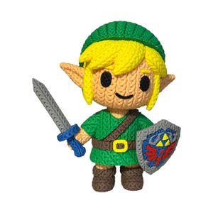 Op de afbeelding: Gebreide figuur van een videogame-personage. De figuur heeft blond haar, een groene tuniek, bruine laarzen en een groene hoed. Het houdt een grijs zwaard en een schild met een rood en blauw embleem vast. Witte achtergrond.