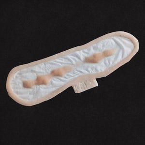 May include: A sanitary pad with a white absorbent core and a pale pink border. The pad has three raised, pinkish-tan areas and a small, rectangular tab. The background is a plain black.