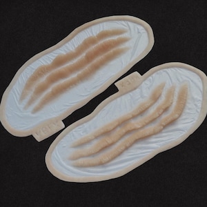 May include: Two prosthetic wound appliances. Each appliance features three raised, flesh-toned lacerations on a white base with a beige border. The text "VBFX" and "X384" are visible on the appliances.