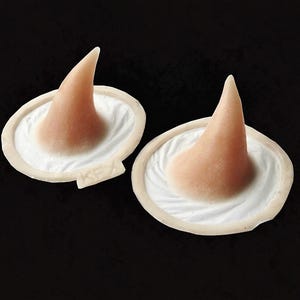 May include: Two prosthetic witch noses in a light skin tone, each mounted on a white base with a ruffled texture. The bases are off-white and have a small rectangular tab. The background is solid black.