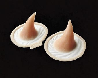 May include: Two prosthetic witch noses, flesh-toned with a pointed tip, sit atop a white, ruffled base. The bases are set against a black background, creating a stark contrast. The noses are likely for Halloween or costume use.