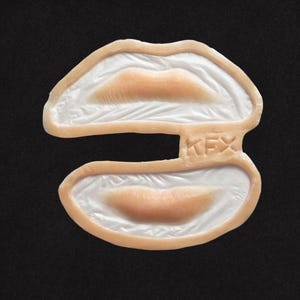 May include: A pair of prosthetic lips, with a light peach-coloured lip shape and a white interior. The lips are separated, with the text "KFX" embossed on the connecting piece. The background is solid black.