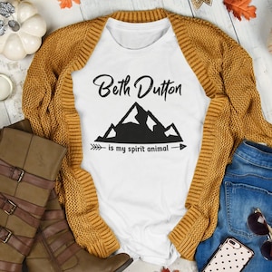 Könnte beinhalten: Weißes T-Shirt mit dem Text "Beth Dutton is my spirit animal" und einer Grafik mit einer Bergkette. Das Shirt liegt auf einer Holzoberfläche mit einem braunen Cardigan, einer blauen Jeans, braunen Stiefeln und einem Handy mit einer gepunkteten Hülle.