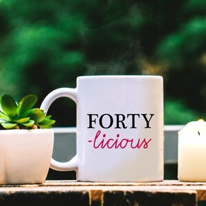Puede incluir: Taza de café blanca con el texto "FORTY -licious" en letras negras y rosas. Una pequeña planta suculenta en maceta y una vela blanca están sobre una superficie de madera.