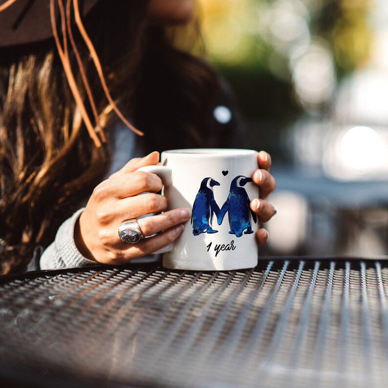 May include: White ceramic mug with a blue watercolor illustration of two penguins holding hands. The mug has the text "1 year" printed on the bottom.
