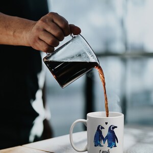 May include: White ceramic coffee mug with a blue and white penguin couple design. The mug has the text "1 year" on the bottom. A hand is pouring coffee from a glass carafe into the mug.