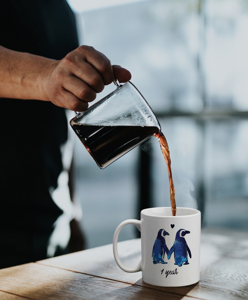May include: White ceramic coffee mug with a blue and white penguin couple design. The mug has the text "1 year" on the bottom. A hand is pouring coffee from a glass carafe into the mug.