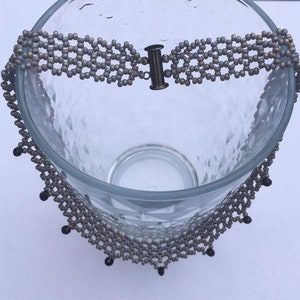 May include: A multi-strand beaded necklace with a silver clasp. The necklace is made of small, iridescent beads in shades of gray and black. The beads are arranged in a geometric pattern, creating a delicate and intricate design.