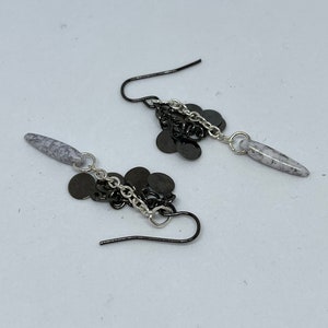 Puede incluir: Un par de pendientes colgantes con una cadena plateada y abalorios de metal negro. Los pendientes tienen una piedra blanca y gris en la parte superior.