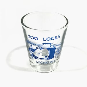 May include: Clear shot glass with a blue graphic of the Soo Locks in Michigan. The text "SOO LOCKS" and "MICHIGAN" are printed in blue on the glass. The shot glass is empty and sits on a white surface.