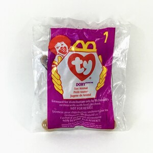 May include: A pink and white Ty Beanie Baby packaging with a brown plush toy inside. The packaging features the Ty logo and the words "DOBY Toy Animal Petit toutor Jugete de Animal". It also includes the text "Licensed for distribution only by McDonald's restaurants with food purchase NOT FOR RESALE".