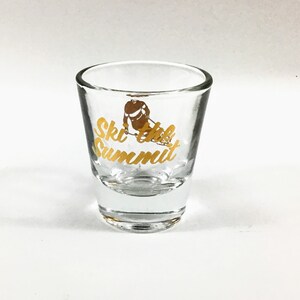 May include: A clear glass shot glass with a gold design that says "Ski the Summit" and a skier in a gold silhouette.