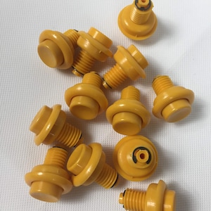 May include: A group of 12 yellow arcade buttons with black stems. The buttons are arranged in a random pattern on a white surface.