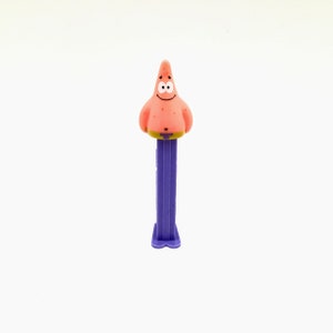 May include: A purple and pink plastic toy with a cartoon character of Patrick Star from Spongebob Squarepants on top. The toy is a dispenser for candy.