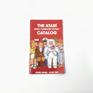 May include: A vintage Atari Video Computer System catalog with a red cover. The cover features the text "THE ATARI VIDEO COMPUTER SYSTEM CATALOG" in white. Below the text is a group of cartoon characters, including an astronaut holding a small American flag. The catalog's tagline is "MORE GAMES. MORE FUN."