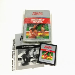 May include: A vintage Atari 2600 game cartridge for RealSports Tennis. The cartridge is black with a silver label that says "Atari 2600" and "RealSports Tennis". The label also features a graphic of two tennis players on a green court.