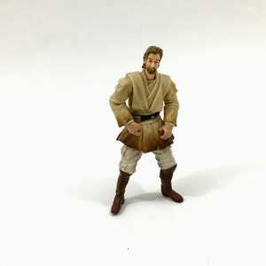 May include: A beige and brown Star Wars action figure of a Jedi Knight with a brown belt and brown boots. The figure has a light brown beard and hair.
