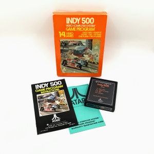 May include: A vintage Atari video game cartridge for the Indy 500 game. The cartridge is black with a green label that reads "game program" and "Indy 500". The cartridge is in its original box, which is orange and features a picture of a race car.