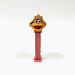 May include: A vintage PEZ dispenser featuring a Sesame Street character with a yellow face, pink lips, and a pink stem. The character has a hat with pink hearts and large, round eyes. The dispenser is on a white background.