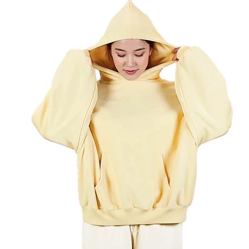 Op de afbeelding: Een lichtgele hoodie met een grote capuchon en een kangoeroezak. De hoodie is gemaakt van een zacht materiaal en heeft lange mouwen. De persoon op de foto draagt de capuchon.