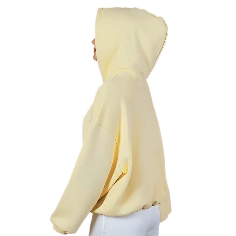 Op de afbeelding: Een lichtgele hoodie met lange mouwen. De hoodie heeft een trekkoord in de taille en een grote capuchon. De persoon draagt een witte broek. De hoodie is gemaakt van een zacht materiaal.