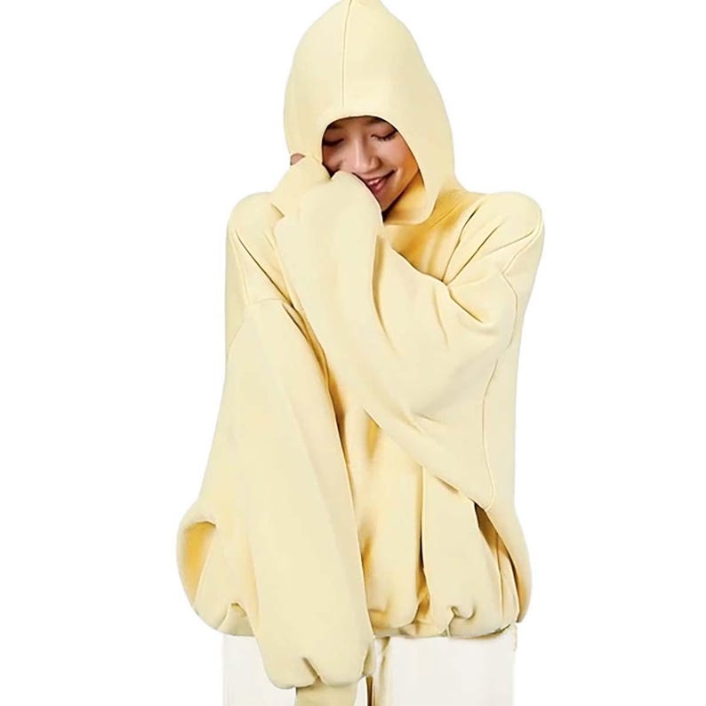 Op de afbeelding: Een lichtgele hoodie met oversized mouwen. De hoodie heeft een grote capuchon en een relaxte pasvorm. De persoon op de foto draagt de hoodie en glimlacht.