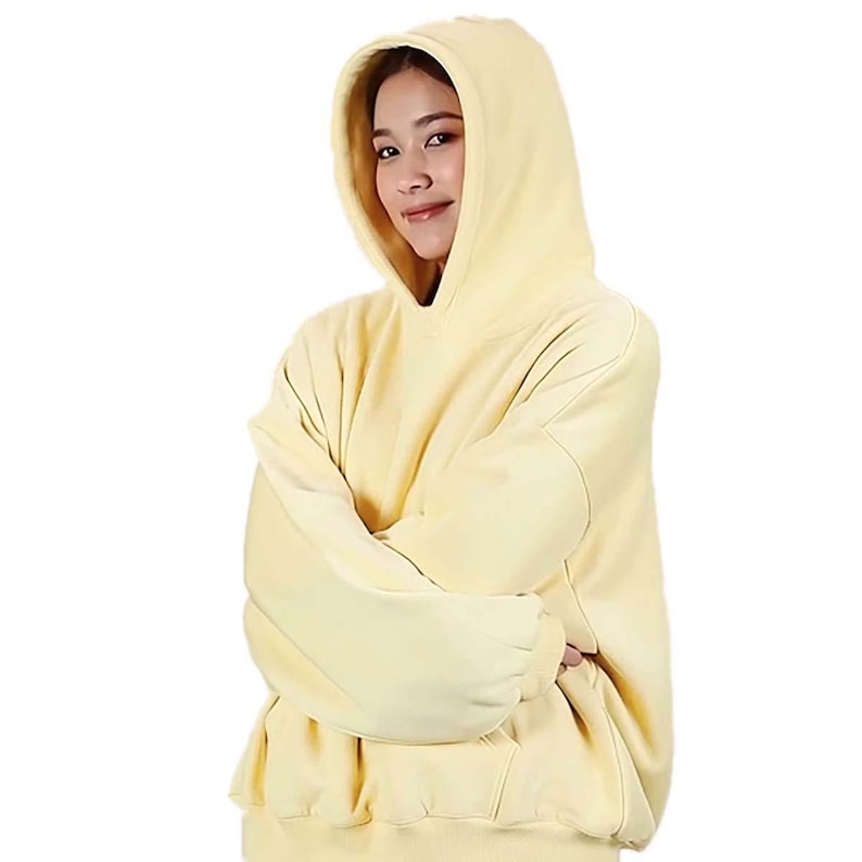 Op de afbeelding: Een lichtgele hoodie met een grote capuchon en lange mouwen. De hoodie is gemaakt van een zacht materiaal en heeft een relaxte pasvorm. De persoon op de foto draagt de hoodie met gekruiste armen.