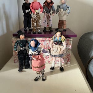 May include: A group of handmade dolls in traditional folk costumes. The dolls are displayed on a purple shelf and a floral box. The dolls have detailed clothing, including hats, aprons, and patterned trousers. The dolls are of various heights and poses.