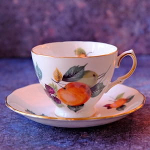 May include: White teacup and saucer set with a gold rim and a fruit design. The teacup features a handle and is decorated with images of peaches, pears, and berries with green leaves. The saucer has a matching fruit design.