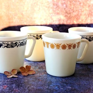 May include: Four white mugs with decorative floral patterns in black and gold. The mugs have a simple, curved handle and are set against a blurred background with a purple and tan color scheme.