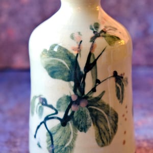 May include: A white ceramic vase with a blue rim and base. The vase is decorated with a floral design featuring green leaves and pink berries. The vase has a narrow neck and a rounded body.