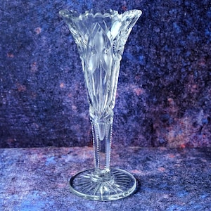 May include: A clear glass vase featuring a flared, scalloped rim and intricate etched floral patterns on the body. The vase stands on a circular, ray-cut pedestal base, with a textured, vertical-ribbed stem connecting the bowl to the base.