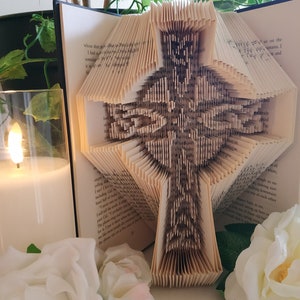 May include: A book sculpture with pages folded into a Celtic cross design. The cross is centered within the open book, with the pages creating a textured, three-dimensional effect. A lit candle and white roses are in the foreground.