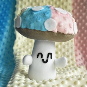 Puede incluir: Un juguete de peluche en forma de hongo con un gorro azul y rosa, lunares blancos y un tallo blanco con una cara sonriente.