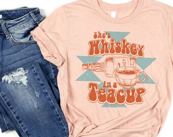 May include: A peach-colored t-shirt with the text "She's Whiskey in a Teacup" in a retro font. The text is surrounded by a blue and orange geometric design. A bottle of whiskey and a teacup are depicted in the design.