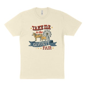 May include: Cream-colored t-shirt featuring a vintage-style graphic. The design includes the text "TAKE ME to the COUNTY FAIR" in red and blue, along with illustrations of a cow, calf, and a Ferris wheel. The shirt has short sleeves.