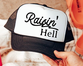 May include: A black and white trucker hat with a white front panel and a black mesh back. The front panel has the text "Raisin' Hell" in a black cursive font.