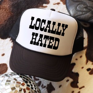 May include: A white and black trucker hat with a black mesh back. The front of the hat has the text "LOCALLY HATED" in black block letters.