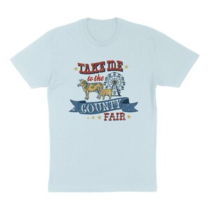 May include: Light blue t-shirt with a distressed graphic. The graphic features the text "TAKE ME to the COUNTY FAIR" in red and blue, along with illustrations of a cow, calf, and a Ferris wheel. The design has a vintage, worn look.