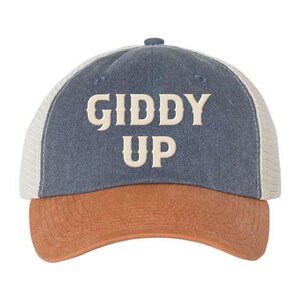 May include: A blue and brown trucker hat with a white mesh back. The hat has the words "Giddy Up" embroidered on the front in a cream color.