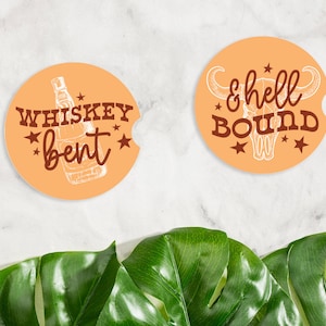 May include: Two orange car coasters with white and brown text. One says "Whiskey Bent" with a whiskey bottle graphic. The other says "& hell BOUND" with a bull skull graphic. Both have star accents.