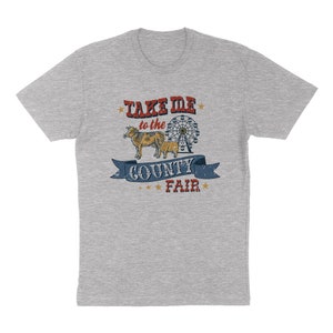May include: A light gray t-shirt featuring a distressed graphic with the text "Take Me to the County Fair." The design includes a Ferris wheel, two cows, and stars. The shirt is a classic crew neck.